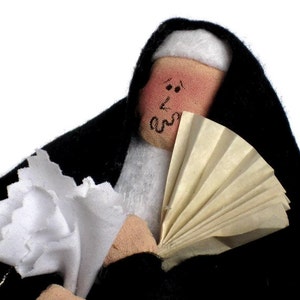 May include: A handmade doll dressed in a black habit and white wimple, holding a white paper fan. The doll has a painted face with rosy cheeks and black eyes.