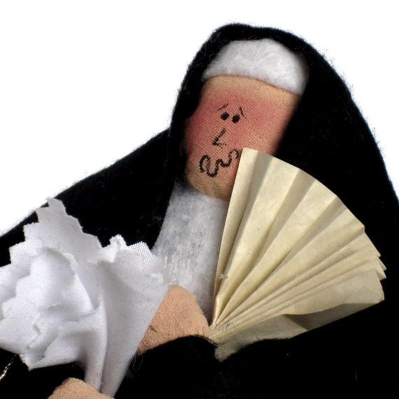 May include: A handmade doll dressed in a black habit and white wimple, holding a white paper fan. The doll has a painted face with rosy cheeks and black eyes.