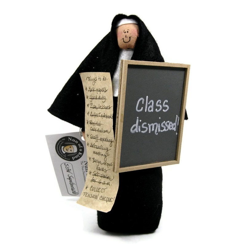 Puede incluir: Una mu&ntilde;eca de fieltro negro vestida de monja sosteniendo una pizarra que dice "&iexcl;Clase terminada!" y una lista de cosas que hacer. La mu&ntilde;eca tiene una cara blanca con una boca sonriente y ojos negros. La mu&ntilde;eca lleva una t&uacute;nica negra y una capucha blanca.