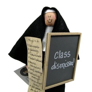 Puede incluir: Una mu&ntilde;eca de monja sosteniendo una pizarra que dice "&iexcl;Clase terminada!" y un rollo con una lista de cosas que hacer.