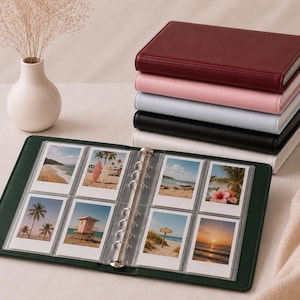May include: A dark green ring-bound photo album open to display eight rectangular instant photos of beach scenes. A stack of five additional photo albums in burgundy, pink, light blue, black, and white sits beside the open album.