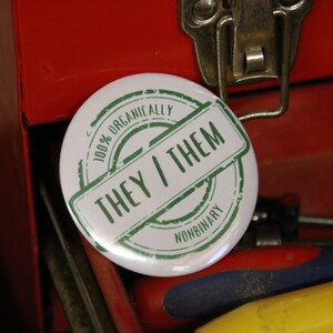 May include: A round white pin-back button featuring a green circular stamp design. The text reads 100% ORGANICALLY, THEY / THEM, and NONBINARY in a bold, sans-serif font. The design mimics a vintage ink stamp with a distressed, weathered texture.