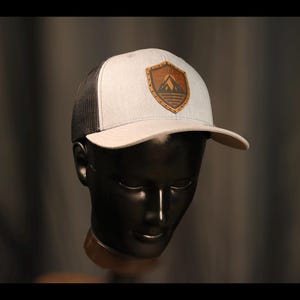 May include: A light gray and black trucker hat featuring a brown leather-like shield patch on the front. The patch displays a mountain range graphic with horizontal lines below it. The hat has a structured front panel and a mesh back.
