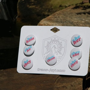 May include: A white card with a mountain logo and the text "trans-joy.com". There are eight round buttons with different pronoun combinations in pink and blue text on a white background. The buttons read "he him", "they he", "he they", "they them", "she her", "they she", "she they", and "like they".