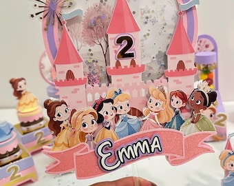 May include: A pink and white princess-themed birthday cake topper with a castle, the number 2, and the name "Emma". The topper features cartoon princesses and a banner. Other princess-themed decorations are in the background.
