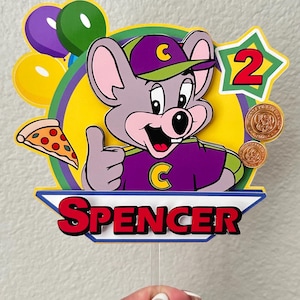 May include: A layered paper cake topper featuring a cartoon mouse character wearing a purple and green cap and shirt, giving a thumbs up. It includes a slice of pizza, balloons, two gold tokens, a star with the number 2, and the name SPENCER in red.