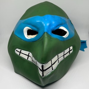 Könnte beinhalten: Grüne und blaue Teenage Mutant Ninja Turtles Maske mit einer weißen Augenmaske und einem weißen und schwarzen Zahndesign.