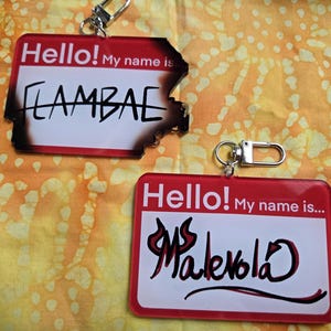 May include: Two red and white name tag keychains with the text "Hello! My name is..." and handwritten names. One says "LAMBAE" with a burnt edge, the other says "Ms. Malevola". Both have silver key rings.
