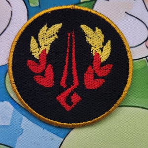 Könnte beinhalten: Runder, schwarz gestickter Aufnäher mit goldenem Rand. Der Aufnäher hat ein rotes und gelbes Design in der Mitte. Das Design umfasst ein zentrales vertikales Element und zwei stilisierte blattartige Formen.