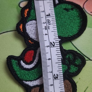 May include: Embroidered Yoshi patch, a green dinosaur character, with a black border and details in white, red, and orange. The patch is approximately 10 cm tall. A measuring tape is placed next to the patch.