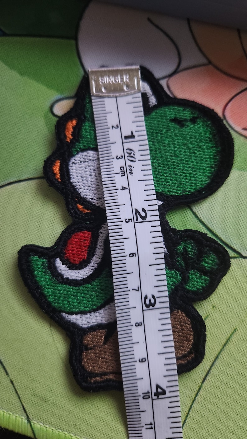 May include: Embroidered Yoshi patch, a green dinosaur character, with a black border and details in white, red, and orange. The patch is approximately 10 cm tall. A measuring tape is placed next to the patch.