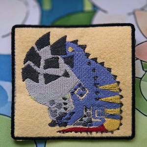 May include: Embroidered patch featuring a blue and yellow dinosaur-like creature with black accents on a tan background. The patch has a black border and is square-shaped. The design is detailed and appears to be machine-embroidered.