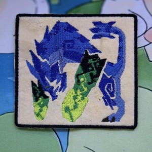 May include: Embroidered patch featuring a blue and green stylized creature design on a cream-colored background. The patch has a black border and is square-shaped. The creature has a long neck and tail.
