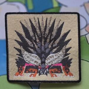 May include: Square embroidered patch featuring a stylized monster face with black, gray, and orange details. The patch has a tan background with a black border. The monster has sharp teeth and a spiky mane.