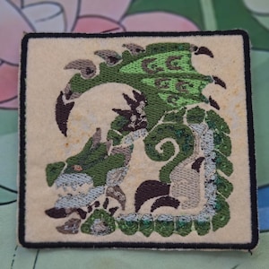 May include: Square embroidered patch featuring a green and brown dragon design on a cream-colored background. The dragon has intricate details and is outlined with a black border. The patch is likely for clothing or accessories.