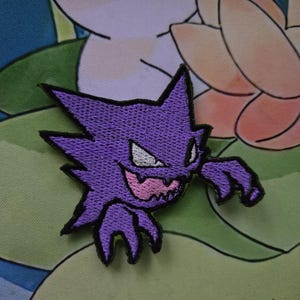 May include: Embroidered purple patch featuring a cartoon ghost-like character with sharp teeth and claws. The patch has a black border and white eyes. The background is a floral pattern with green and white colors.