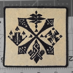 May include: Square patch with a cream-colored background and a black geometric design. The design features a central cross shape with ornate elements at each end. The patch is bordered with a black stitched edge.