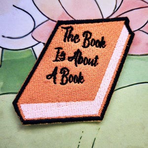 Op de afbeelding: Geborduurde patch in de vorm van een boek met de tekst "The Book Is About A Book" in zwart schrift op een oranje achtergrond. Het boek heeft een witte rug en een zwarte rand. De patch staat op een bloemenachtergrond.