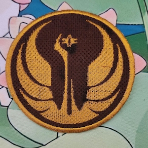 May include: Circular embroidered patch with a brown and gold emblem. The design features stylized wings and a central symbol, all outlined with gold stitching. Suitable for application on clothing or accessories.