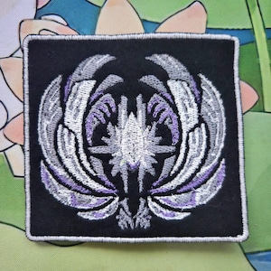 May include: Square black embroidered patch featuring a symmetrical design in white, gray, and purple. The design appears to be a stylized emblem or crest with wing-like elements. The patch has a white border.