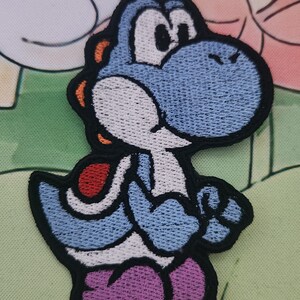 May include: Embroidered patch of a blue Yoshi character with a white belly and purple shoes. The patch has a black border and orange accents. The background features a light green and pink floral design.