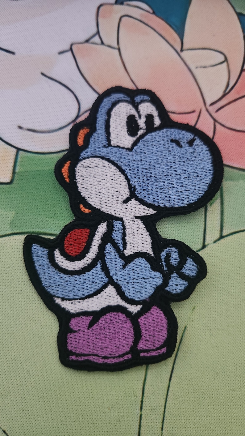 May include: Embroidered patch of a blue Yoshi character with a white belly and purple shoes. The patch has a black border and orange accents. The background features a light green and pink floral design.