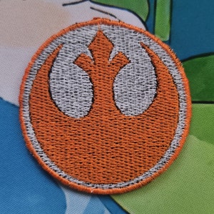 May include: Embroidered patch depicting the Rebel Alliance symbol. The patch is circular, with an orange outer ring and a white and orange emblem in the centre. The patch is on a colourful background.