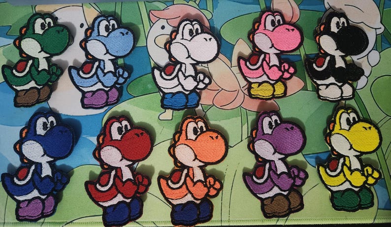 May include: A selection of embroidered Yoshi patches from the Super Mario series. The patches depict Yoshi in a range of colours: green, blue, pink, black, yellow, red, orange, and purple. Each patch is intricately designed and suitable for clothing or accessories.