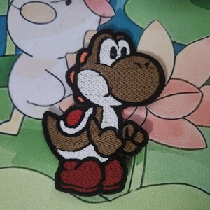 May include: Embroidered patch of a brown Yoshi character with a white belly, red shoes, and orange details. The patch has a black border and is designed for clothing or accessories. The design is based on a popular video game character.
