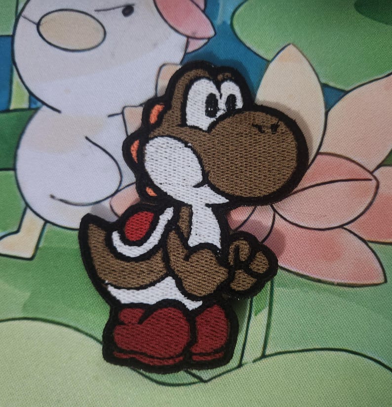 May include: Embroidered patch of a brown Yoshi character with a white belly, red shoes, and orange details. The patch has a black border and is designed for clothing or accessories. The design is based on a popular video game character.