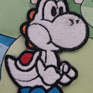 May include: Embroidered patch of Yoshi, a white character with black outlines, orange details, a red saddle, and blue boots. This patch is designed for application on clothing or accessories, featuring a well-known video game figure.