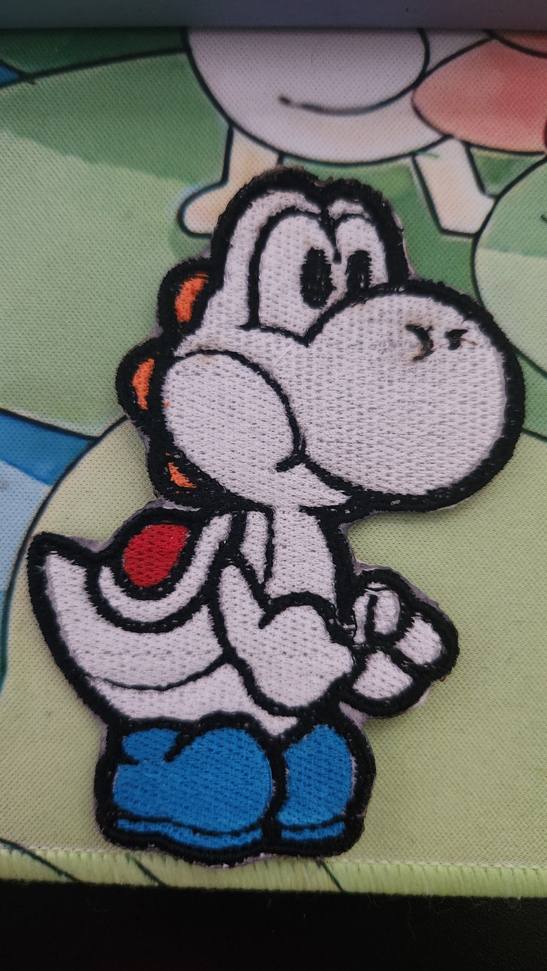 May include: Embroidered patch of Yoshi, a white character with black outlines, orange details, a red saddle, and blue boots. This patch is designed for application on clothing or accessories, featuring a well-known video game figure.