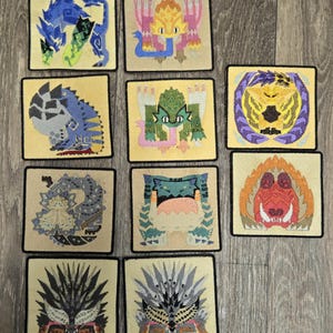 Könnte beinhalten: Eine Sammlung von zehn quadratischen, gestickten Aufnähern mit verschiedenen Monsterdesigns. Jeder Aufnäher hat einen schwarzen Rand und einen beigen Hintergrund. Die Monster sind in verschiedenen Farben dargestellt, darunter Blau, Grün, Gelb und Orange.
