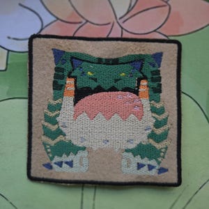 May include: Embroidered patch featuring a green monster with blue accents, a pink tongue, and orange details. The patch is square with a tan background and a black border. The design is detailed and colorful.