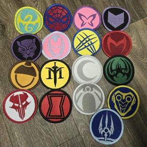 May include: A collection of round embroidered patches featuring various superhero symbols. The patches are in a variety of colors, including blue, red, pink, and yellow. The designs include a spider web, a butterfly, and a crescent moon.