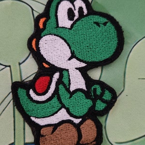 May include: Embroidered patch of Yoshi, a green dinosaur character from the Super Mario series. The patch has a black border, white belly, brown shoes, and red saddle. The patch is approximately 7.6 cm tall.