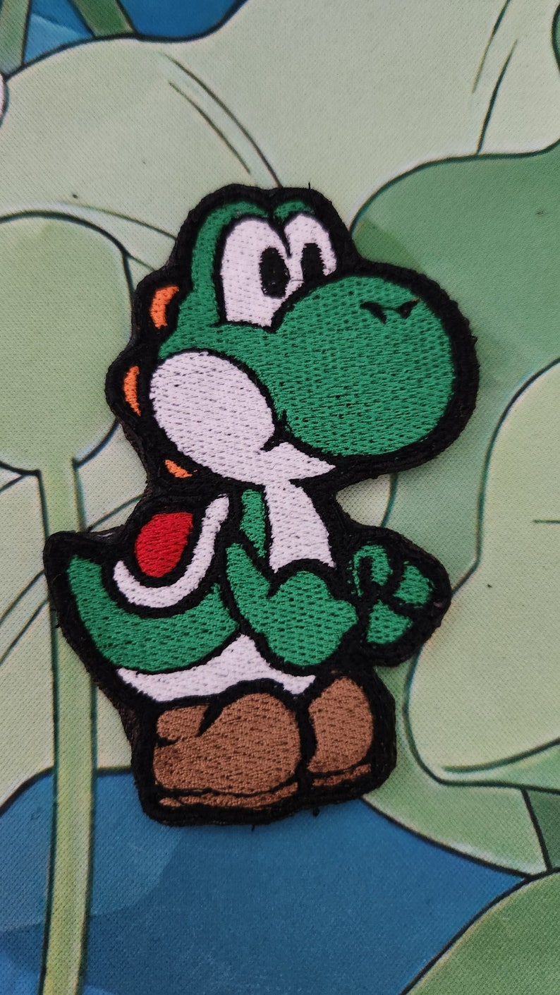 May include: Embroidered patch of Yoshi, a green dinosaur character from the Super Mario series. The patch has a black border, white belly, brown shoes, and red saddle. The patch is approximately 7.6 cm tall.