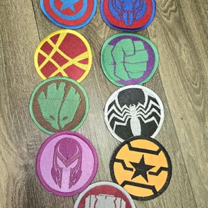 Op de afbeelding: Een verzameling ronde geborduurde patches met verschillende superheldensymbolen. De patches bevatten een ster, een spin, een vuist en andere iconische ontwerpen in kleuren als rood, blauw, groen en geel. Ze zijn gerangschikt op een houten oppervlak.