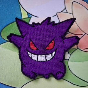 Könnte beinhalten: Gestickter lila Aufnäher mit einem Cartoon-Geist mit roten Augen und einem breiten, zahnigen Grinsen. Der Aufnäher hat einen stacheligen Kopf und ausgestreckte Arme. Das Design befindet sich auf einem hellgrünen und blauen Hintergrund.