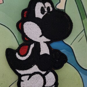 May include: Embroidered patch of a black Yoshi character with white and red detailing. The patch is designed in the shape of the character and is likely for application on clothing or accessories. The background features a green and blue floral pattern.