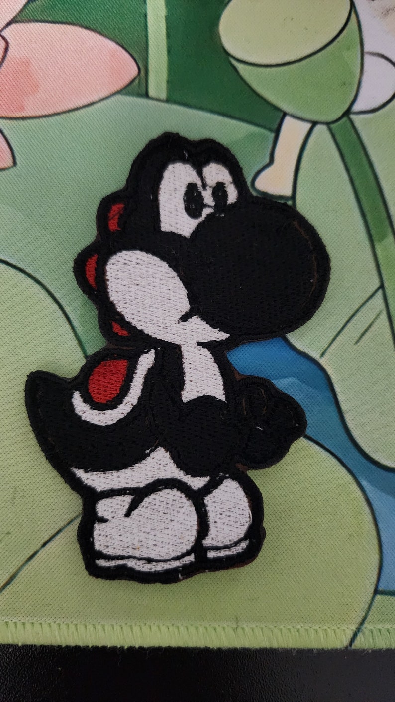 May include: Embroidered patch of a black Yoshi character with white and red detailing. The patch is designed in the shape of the character and is likely for application on clothing or accessories. The background features a green and blue floral pattern.