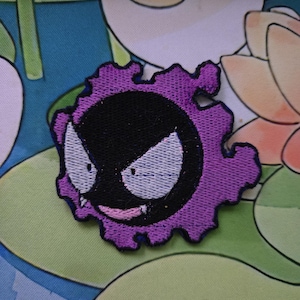 May include: Embroidered patch featuring a purple and black cartoon character with white eyes and a small mouth. The patch has a jagged, scalloped edge. The character is set against a colorful background.