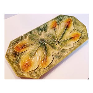 May include: A rectangular ceramic serving platter with a raised design of three overlapping fig leaves. The leaves are in shades of green, yellow, and brown, and the background is a woven green pattern.