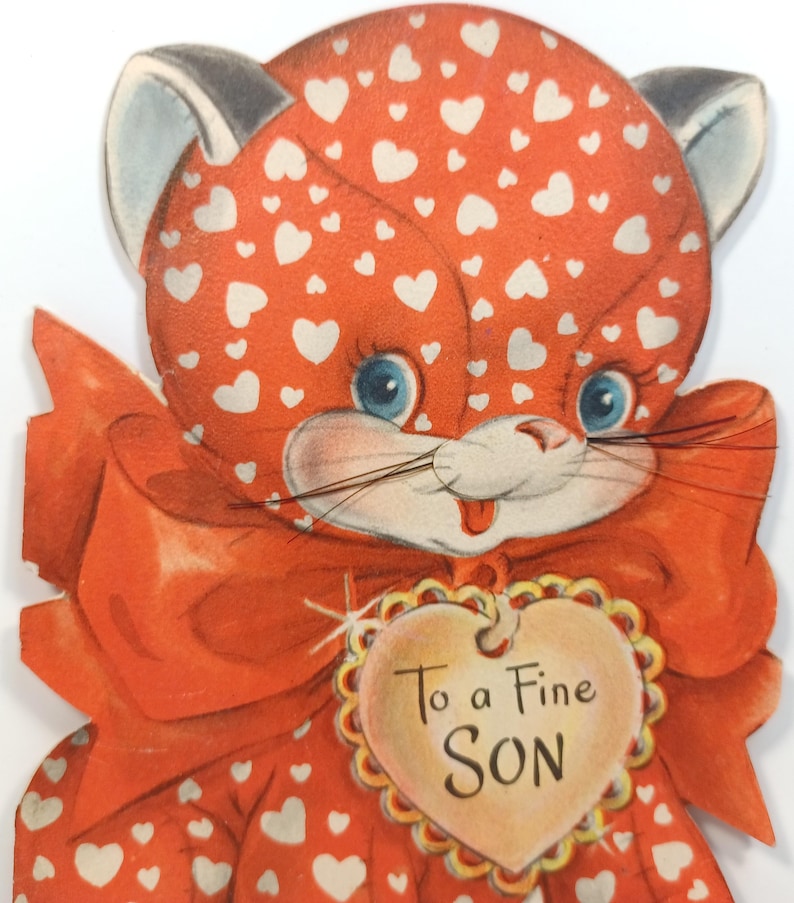 Op de afbeelding: Vintage Valentijnsdagkaart met een cartoonkat met oranje vacht en witte hartpatronen. De kat heeft blauwe ogen, een rode strik en een hartvormig label met de tekst "To a Fine SON".