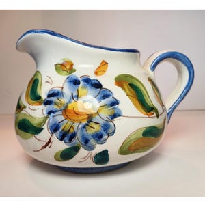 May include: White ceramic pitcher with a blue rim and handle, adorned with a large blue and yellow floral design. Green and orange leaf accents are scattered around the pitcher's body. The pitcher has a spout for pouring.