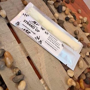 May include: A white and black shampoo packet with the text "STRAND UP" and "eucalyptus orange" on a wooden surface. The packet contains a bar of shampoo. The background includes small stones.