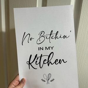 Puede incluir: Un cartel blanco con texto negro que dice "No Bitchin' in my Kitchen". El cartel tiene una pequeña ilustración en blanco y negro de una espátula y un batidor cruzados con hojas debajo del texto.