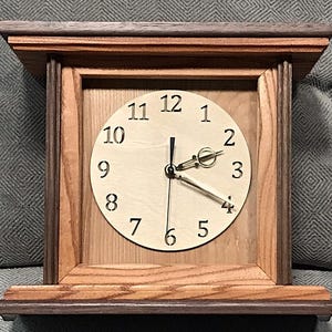 May include: A wooden mantel clock with a light-colored face and black numerals. The clock has a rectangular frame with a dark brown top and base. The clock hands are gold-colored.