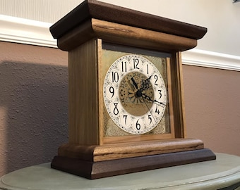 Può includere: Un orologio da mensola in legno con corpo in rovere chiaro e parte superiore e base marrone scuro. Il quadrante dell'orologio è bianco con numeri neri e dettagli ornati. Le lancette dell'orologio sono nere.