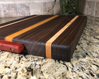 Puede incluir: Tabla de cortar rectangular de madera con superficie marrón oscuro y rayas de madera más claras. Un asa roja está fijada al lateral. La tabla mide aproximadamente 30 cm de largo.